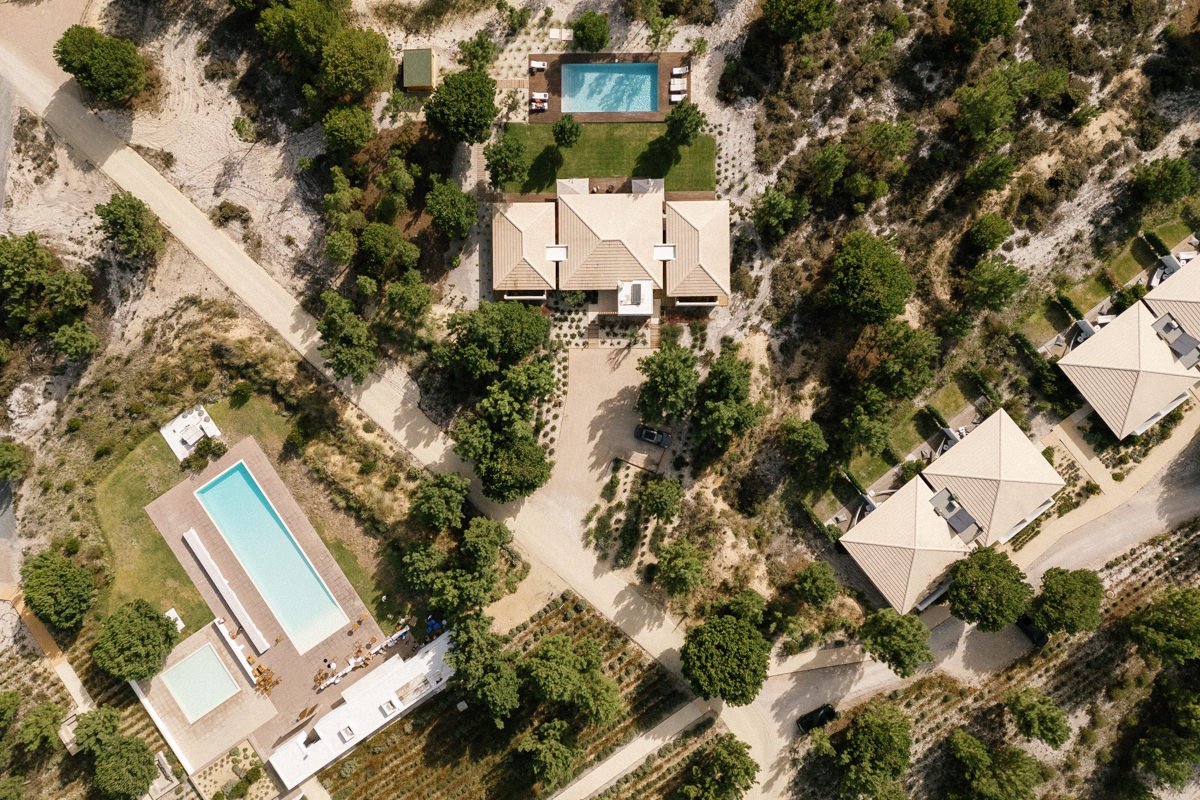 Aerial view of a landscaped residential at Spatia Comporta in a Destination Wedding area with several houses, green trees, and two rectangular swimming pools. Dirt roads and pathways connect the properties, which are surrounded by sand and natural vegetation.