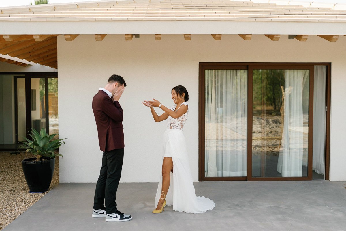 A bride in a white dress excitedly gestures toward her groom in a burgundy jacket and black pants, who covers his face in emotion during their Portugal wedding at Spatia Comporta. They stand on a patio outside a modern building with large windows.