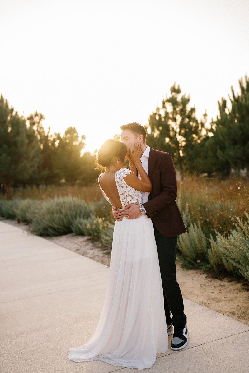 A bride in a white dress and a groom in a burgundy jacket share a kiss outdoors at sunset, standing on a path surrounded by greenery, beautifully captured by photographer Arte Magna at their Portugal Destination wedding.